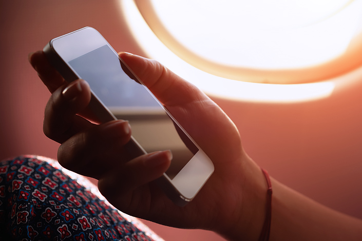 A woman sitting in an aircraft cabin holding a mobile phone in her hand with the "Airplane / Flight Safe" mode activated